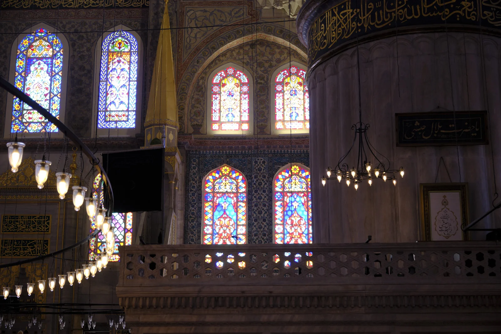 Stained-glass windows and a stone balcony inside the Blue Mosque, with hanging lamps and a column banded in gold script.