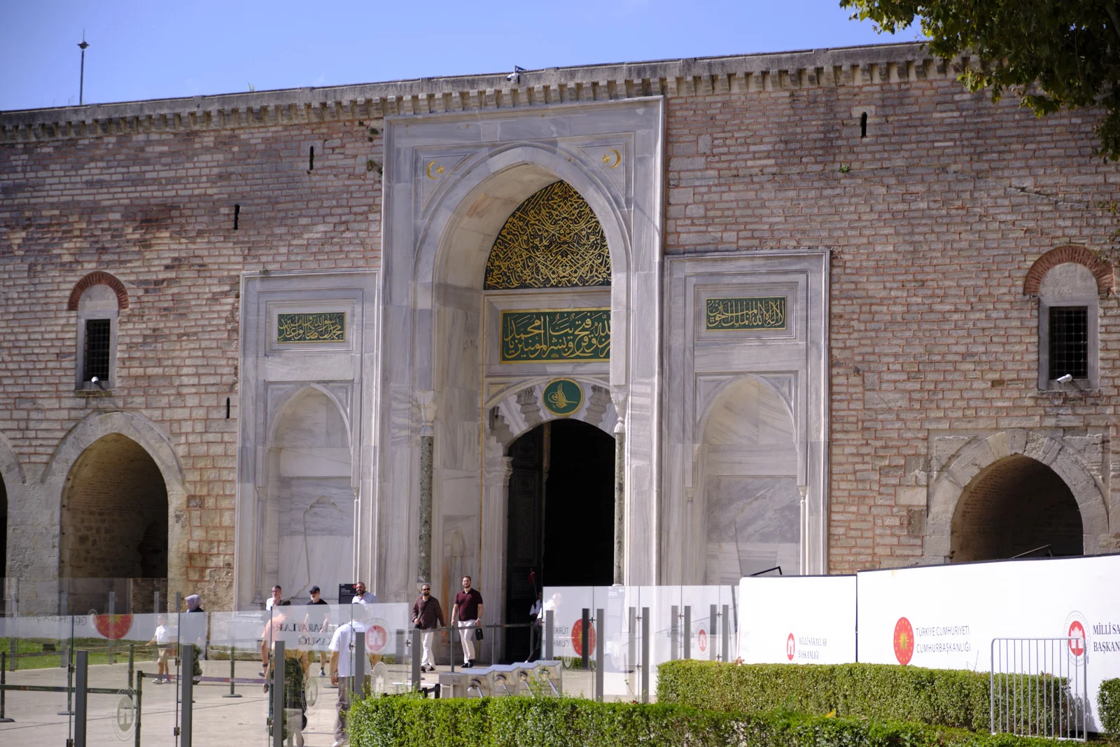 The marble Imperial Gate of Topkapı Palace, with gold calligraphy in the arch, set in a brick wall behind barriers and hedges.