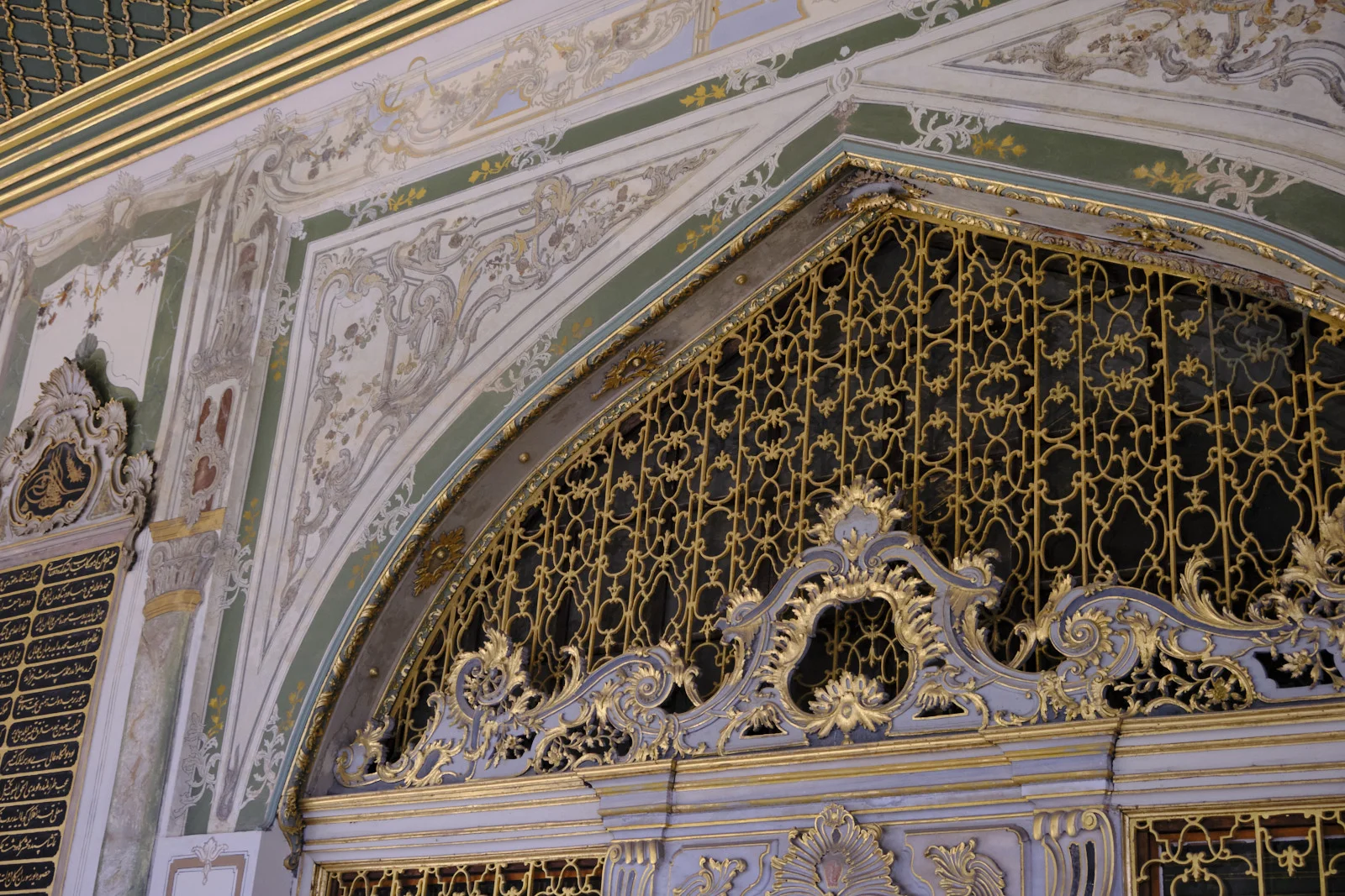 An ornate gold iron grille fills a painted rococo arch, with scrolling gold decoration and a tughra roundel.