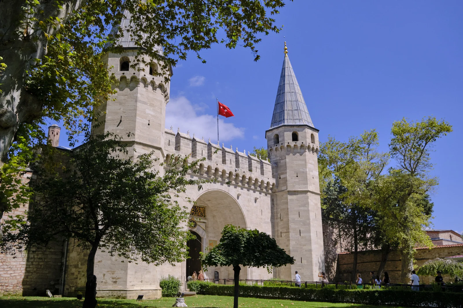 The twin-towered Gate of Salutation at Topkapı Palace, with a Turkish flag above the arch and visitors on the path under a blue sky.