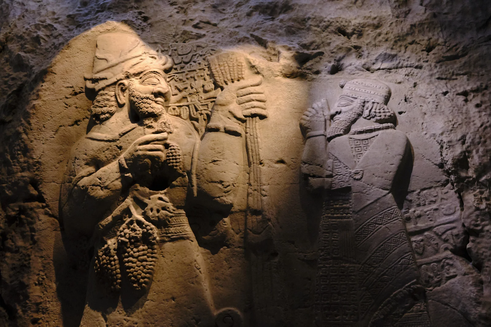 A museum spotlight on an ancient stone relief of two bearded men, one in a tall headdress and one in a patterned robe.