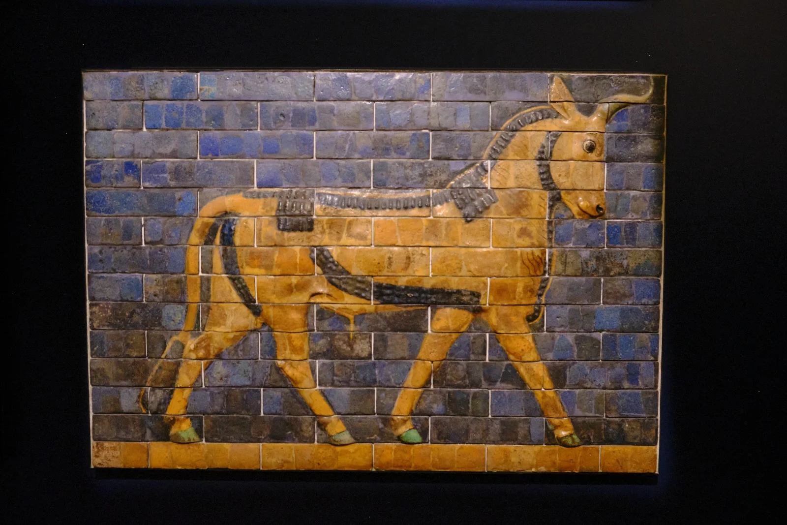 A glazed-brick relief of a yellow bull striding across blue tiles, displayed on a black museum wall.