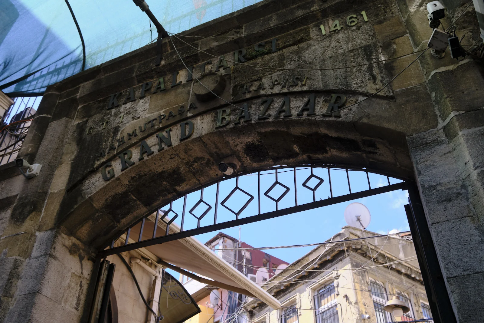 A weathered stone arch lettered Grand Bazaar, with an open iron gate looking onto buildings under a blue sky.