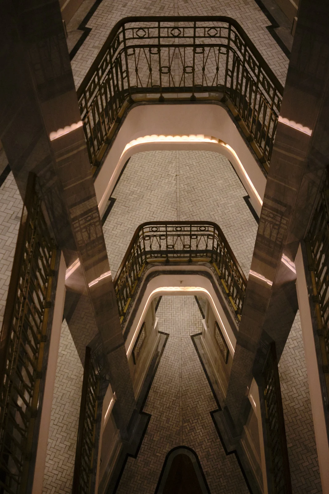 A top-down view of a stairwell with herringbone tile floors, iron railings, and warm light along each landing.