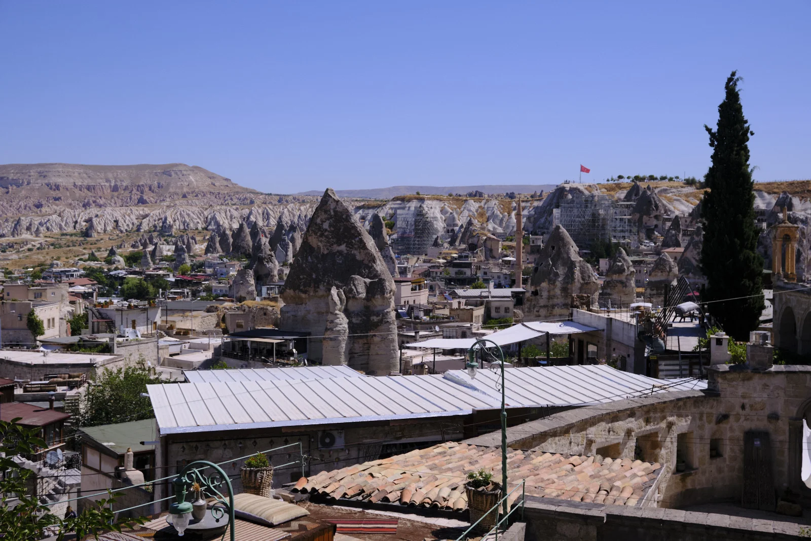 A sunny view over Göreme with terracotta and white metal roofs in front, conical fairy chimneys and a minaret among the houses, and eroded white hills beyond.