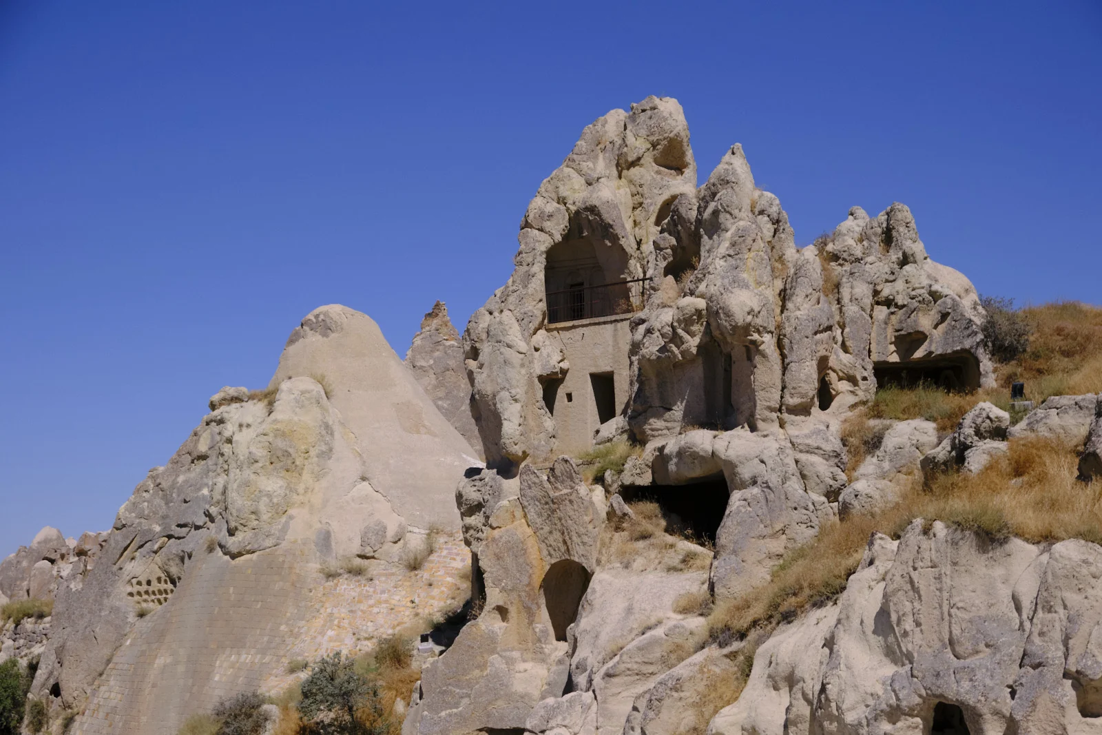 Jagged pale rock with carved rooms, a barred window, and a large conical outcrop under a deep blue sky.