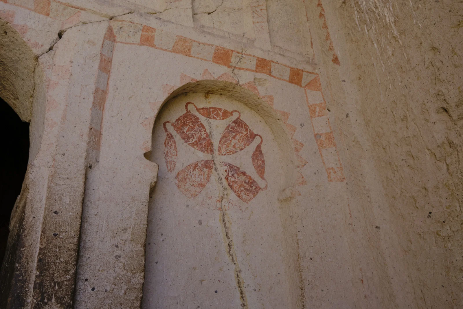A worn red-ochre cross is painted inside an arched niche on cracked plaster, with a checkerboard stripe and a zigzag border above it.