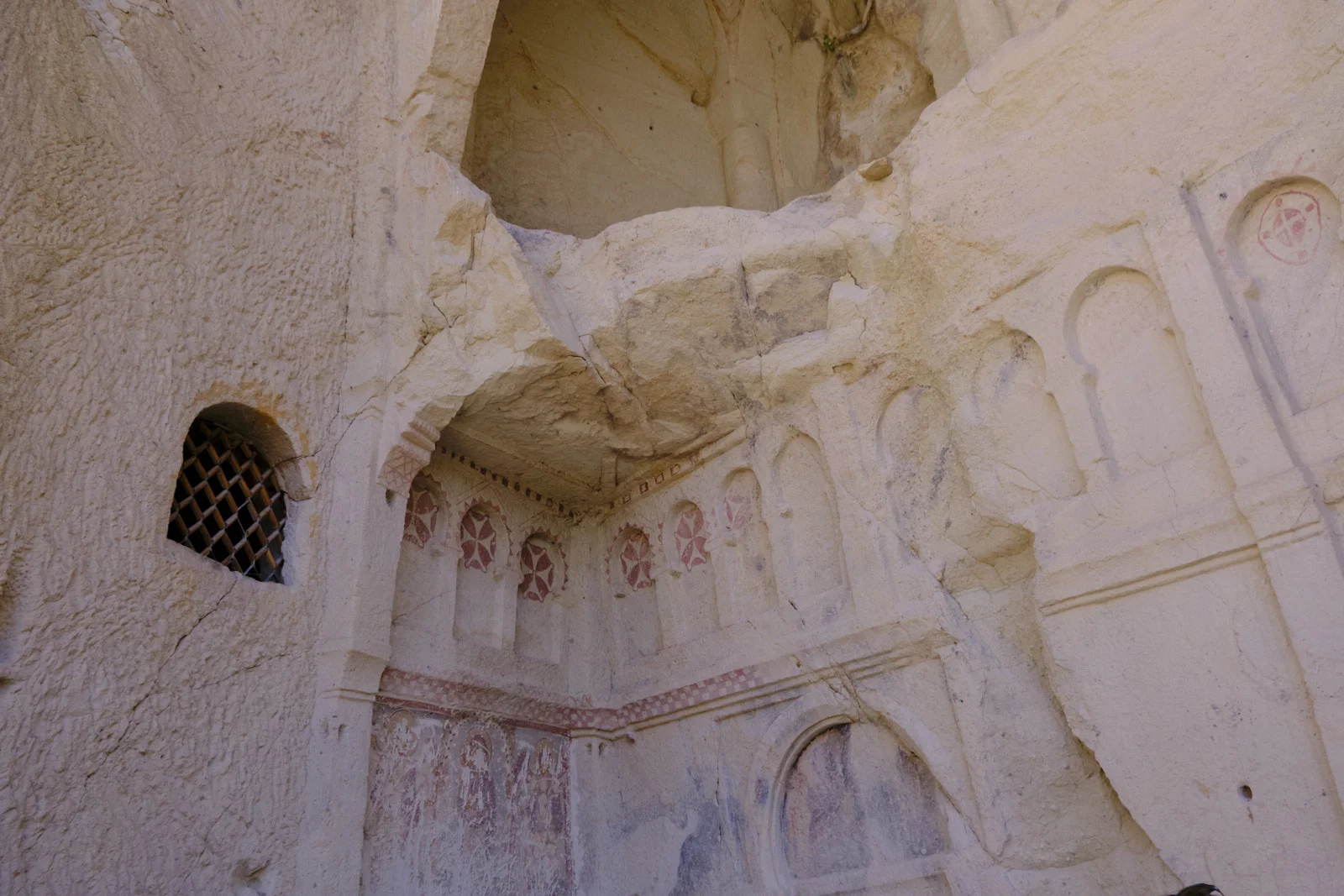 Rock-cut church interior with a grated window, worn frescoes of standing figures, red crosses in arched niches, and a collapsed section of ceiling.