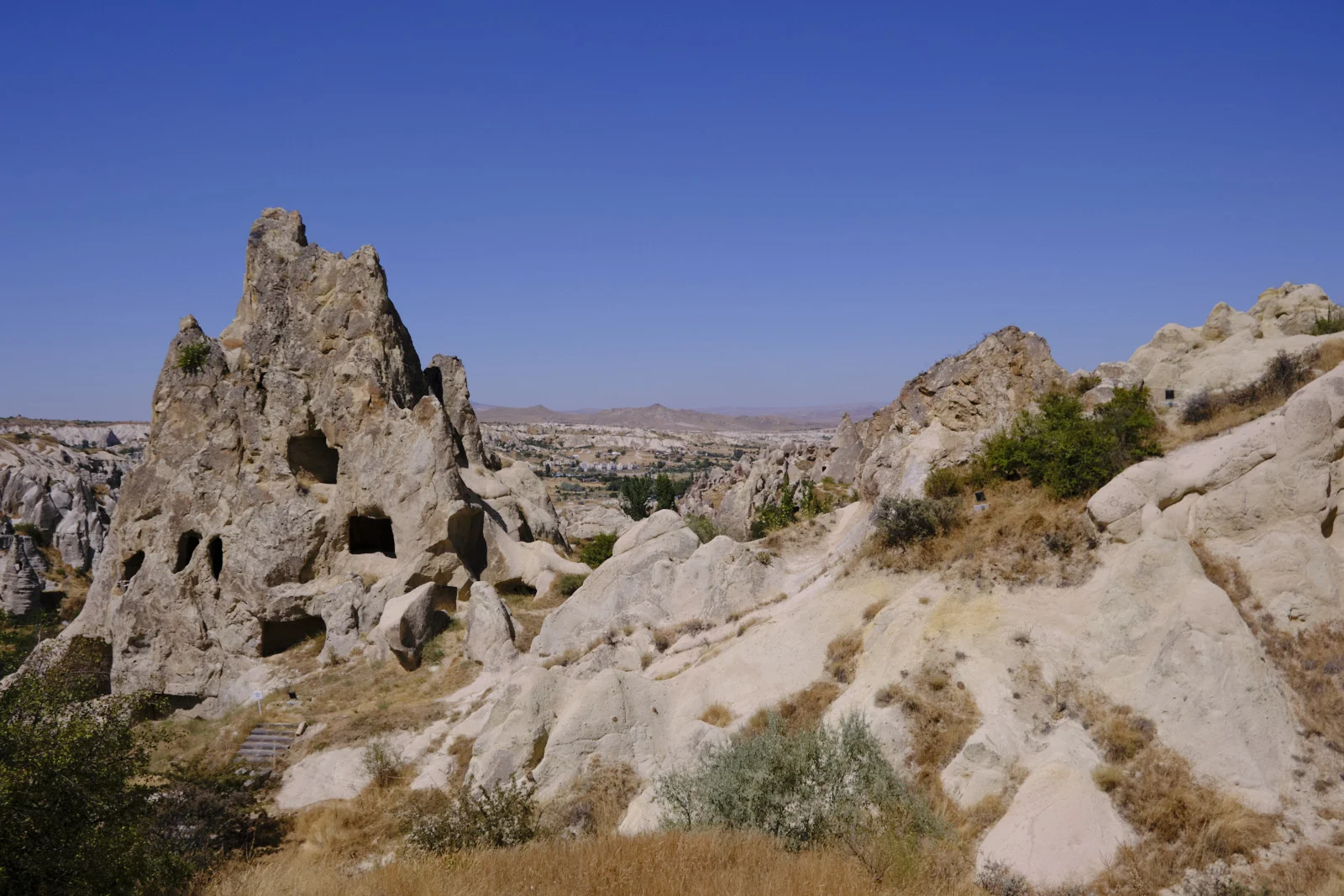 A tall rock pinnacle with cave openings overlooks a wide valley of pale stone, dry grass, and a distant town under a clear sky.