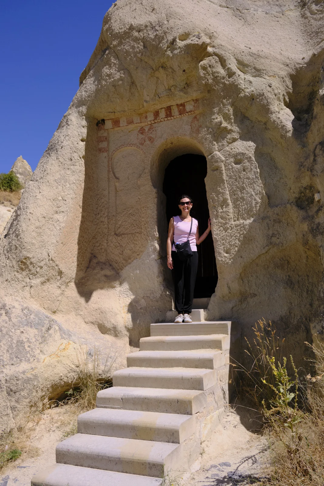 Concrete steps lead up to a woman standing in a rock-cut church doorway decorated with faded red geometric painting.