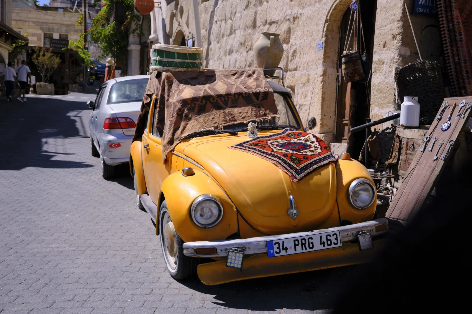 A yellow vintage Beetle covered with patterned rugs is parked on a cobblestone street beside stone shopfronts and hanging carpets.