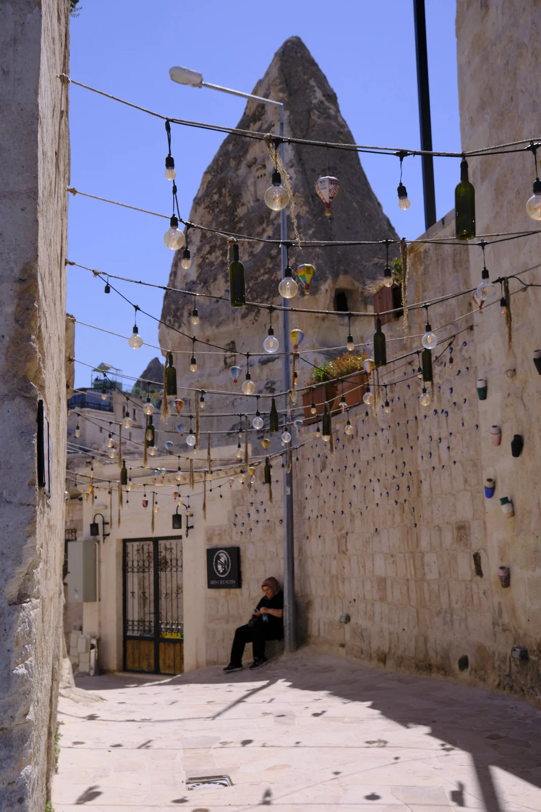 A narrow stone lane hung with light bulbs and green bottles, a woman seated by a gate, and a tall fairy chimney against a blue sky.