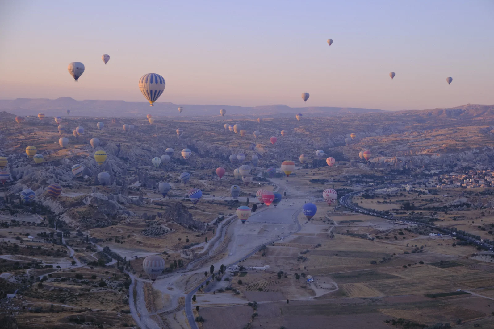 Many colorful hot-air balloons float over a wide eroded valley at dawn, with roads, fields, and a town visible below.