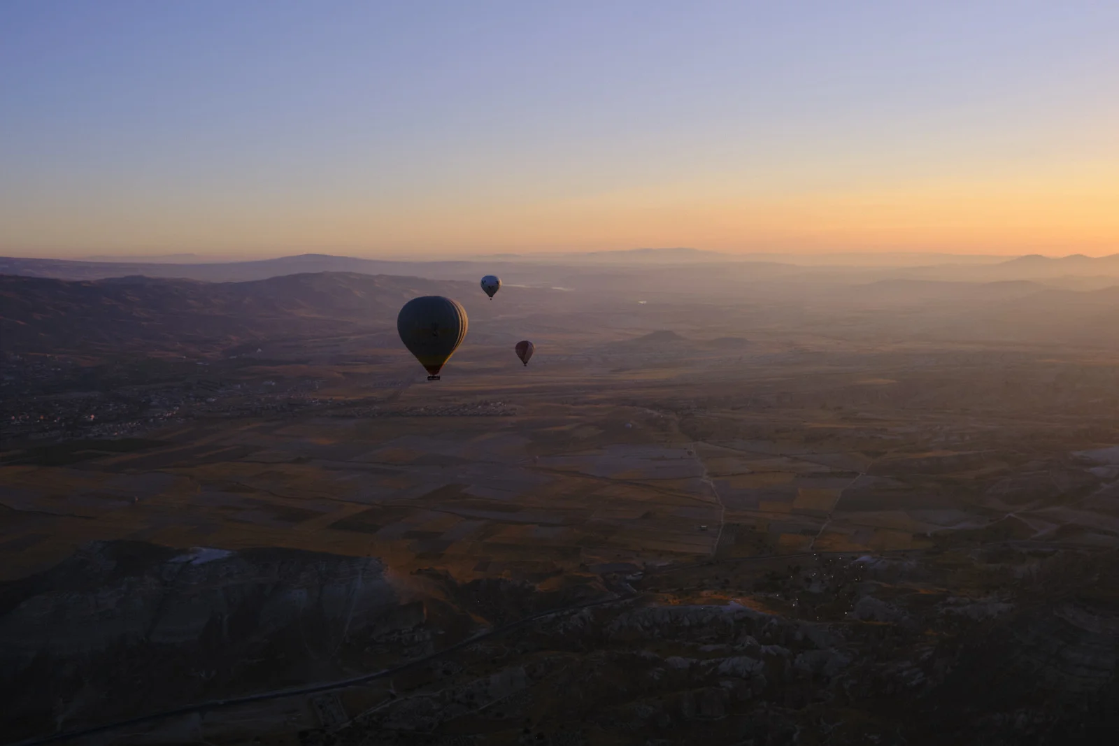 Three hot-air balloons float over brown and gold farmland at sunrise, with hazy mountain ridges along the horizon.