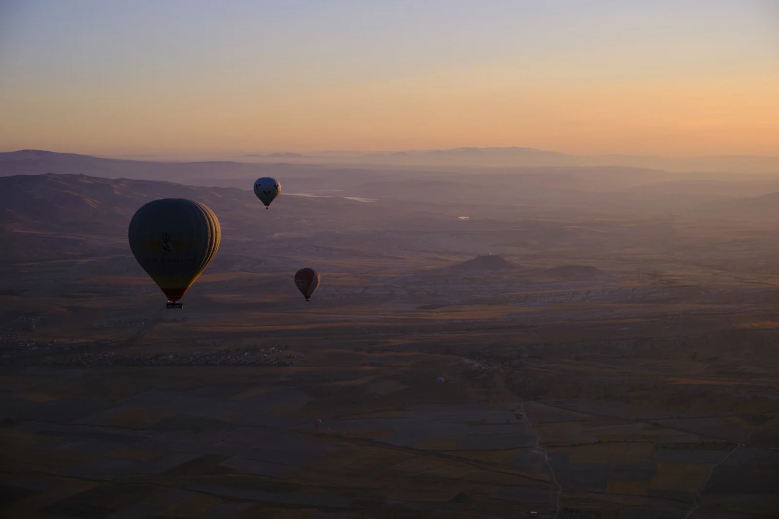 A large hot-air balloon in the left foreground and smaller balloons beyond it, above a wide patchwork of fields at sunrise.
