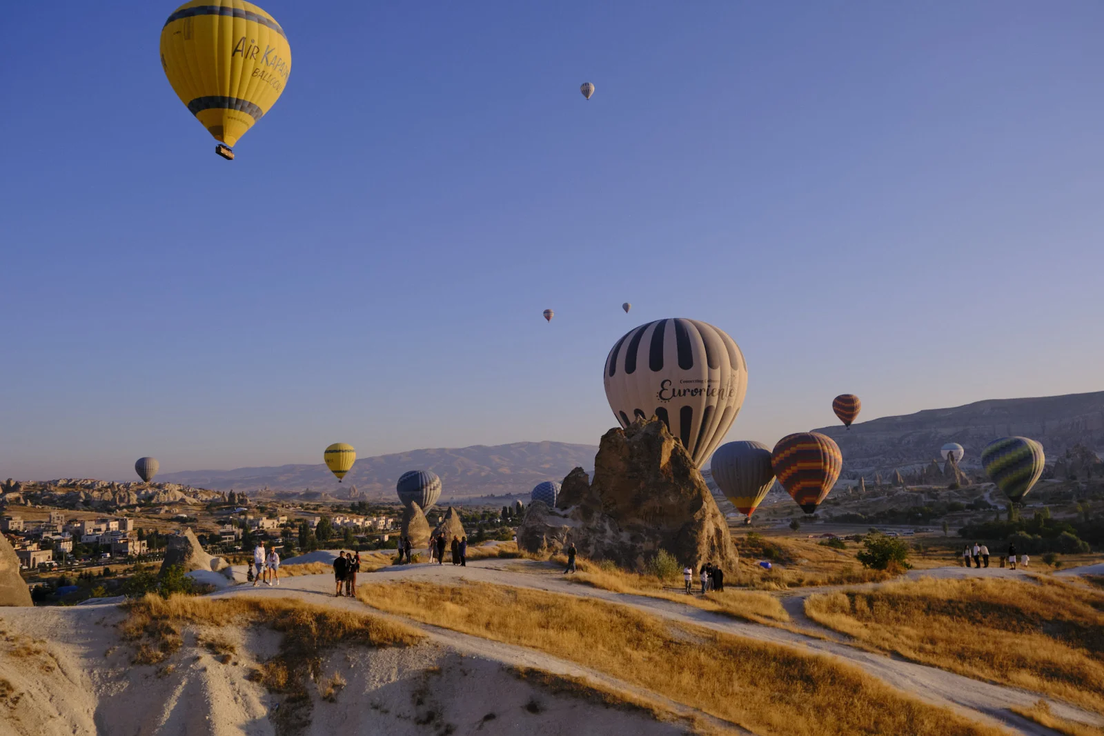 People on a dirt path along a rocky ridge watch hot-air balloons, including a large yellow one and a striped balloon beside a rock pinnacle.