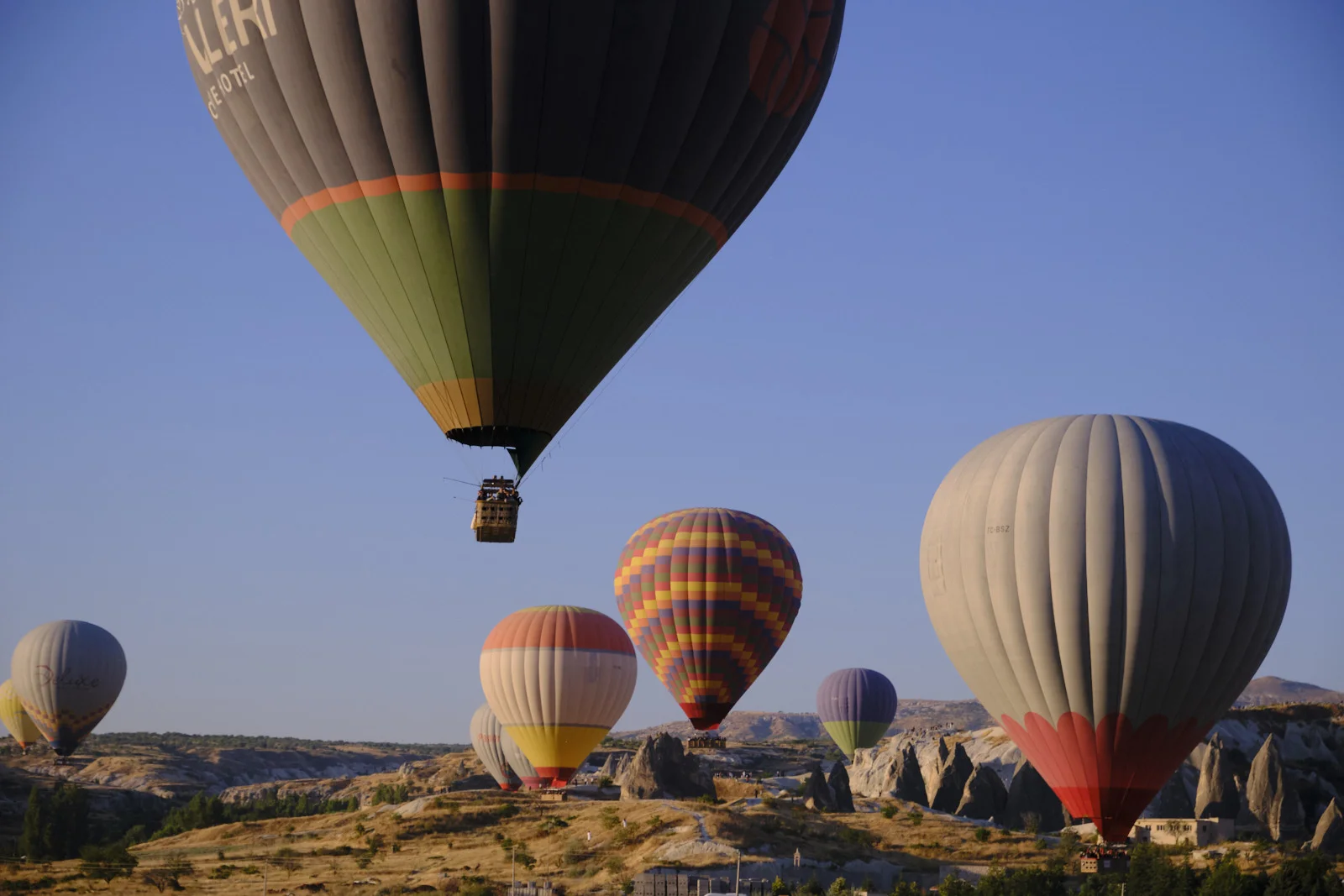 A huge hot-air balloon and its passenger basket pass overhead, with more balloons and rock pinnacles in the valley below.