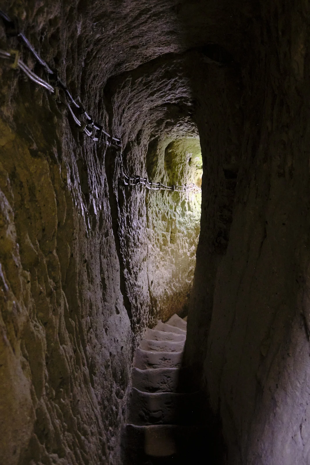 A dim volcanic-rock tunnel with rough steps and a metal chain along the wall, opening toward bright light.