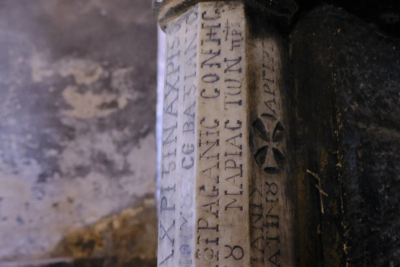 Close view of a stone column inscribed with Greek letters and a carved cross, beside a dark worn surface.
