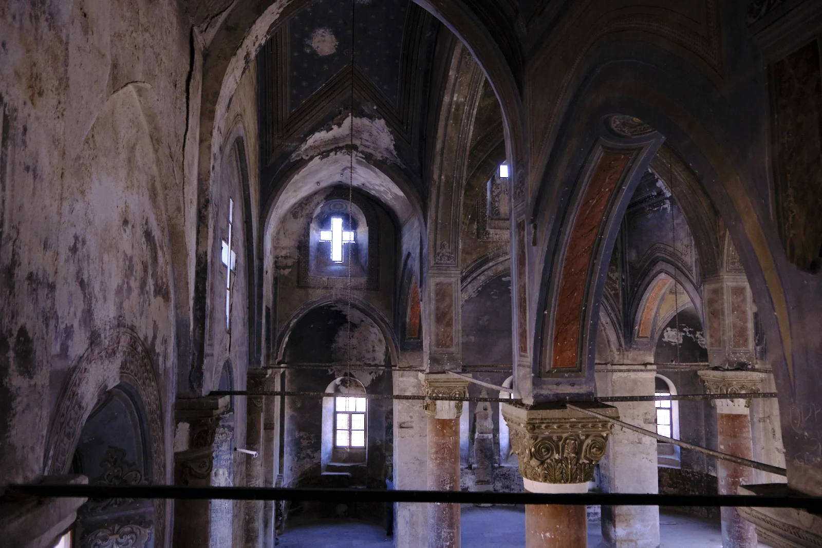 A tall church nave with peeling plaster, painted vaults, stone columns, and a bright cross-shaped window at the far end.