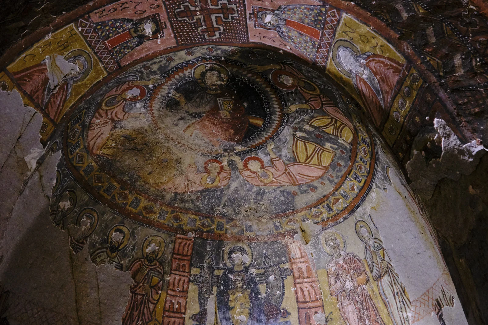 A worn apse fresco with Christ seated in a dark circular mandorla, surrounded by figures, and standing saints painted beneath a band of ornament.