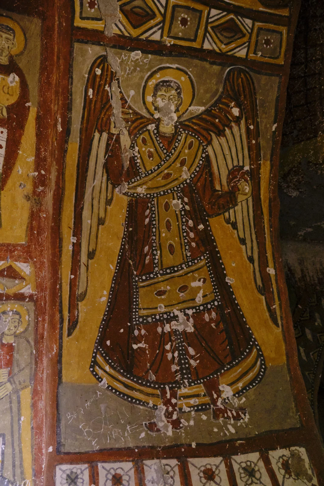 A weathered fresco of a standing archangel in a red and gold robe with a large yellow halo, spread wings, and an orb in one hand.