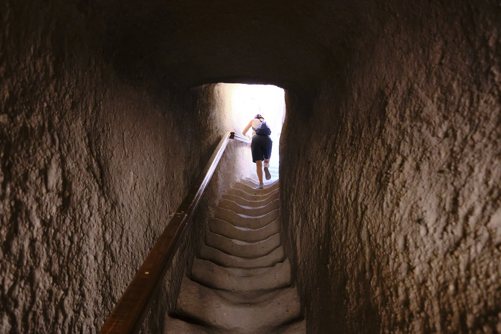 A woman with a backpack climbs steep stone steps inside a narrow rock-cut stairwell toward a bright opening.