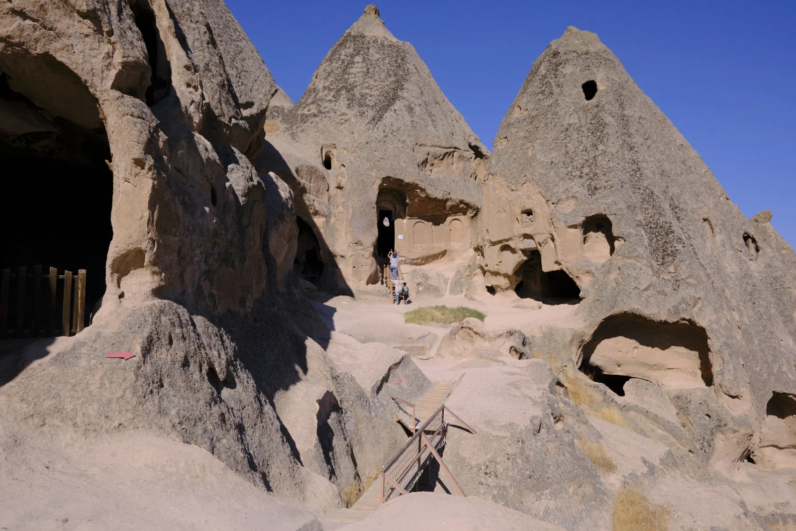 Sunlit conical rocks riddled with cave openings, a person in a carved doorway, and a metal staircase in the foreground under a deep blue sky.