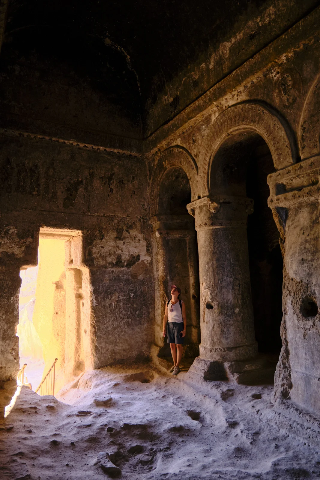 A woman in a cap and backpack stands on a sandy floor looking up at massive carved columns and arches in a dim rock-cut hall, with bright light from a doorway.