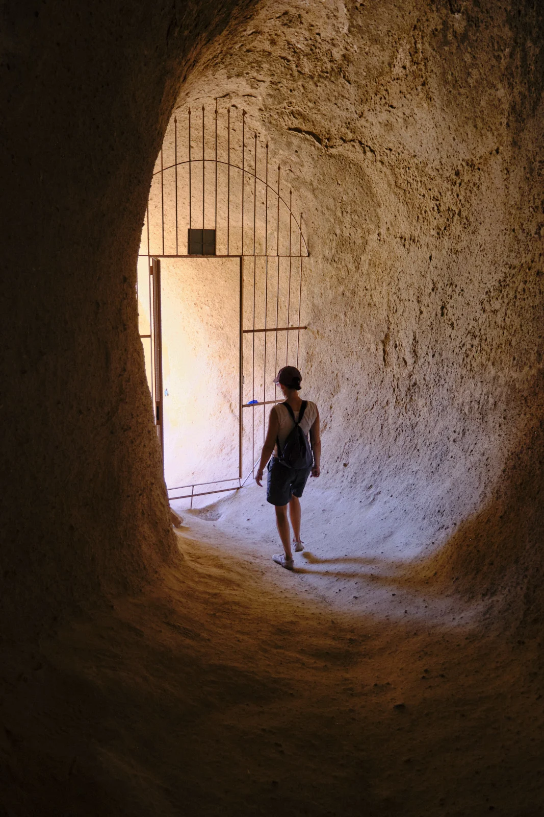 A woman walks away through a curved rock tunnel toward a tall iron gate standing open in bright light.