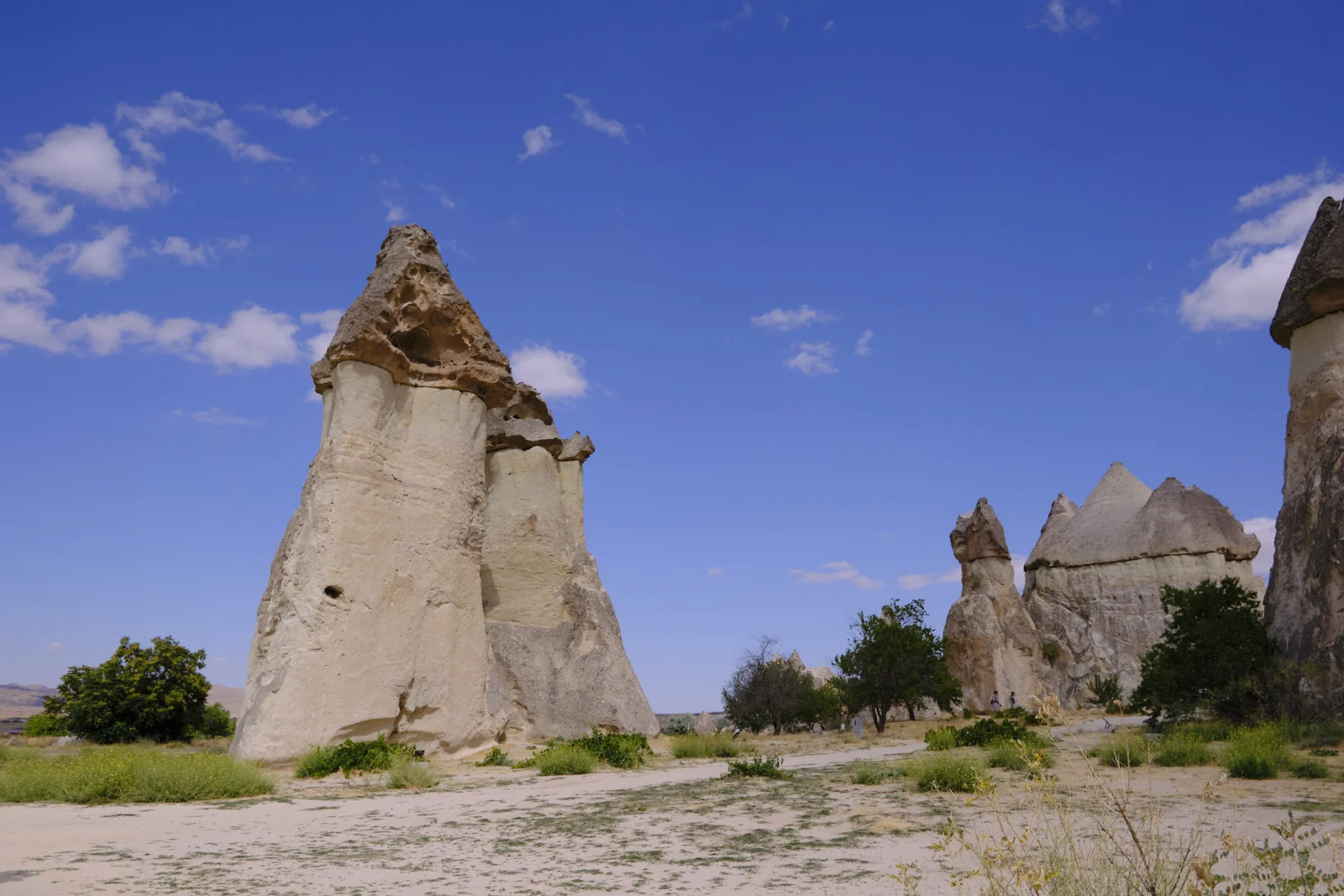 Tall cream-colored fairy chimneys with eroded caps stand beside a sandy path under a blue sky with scattered clouds.