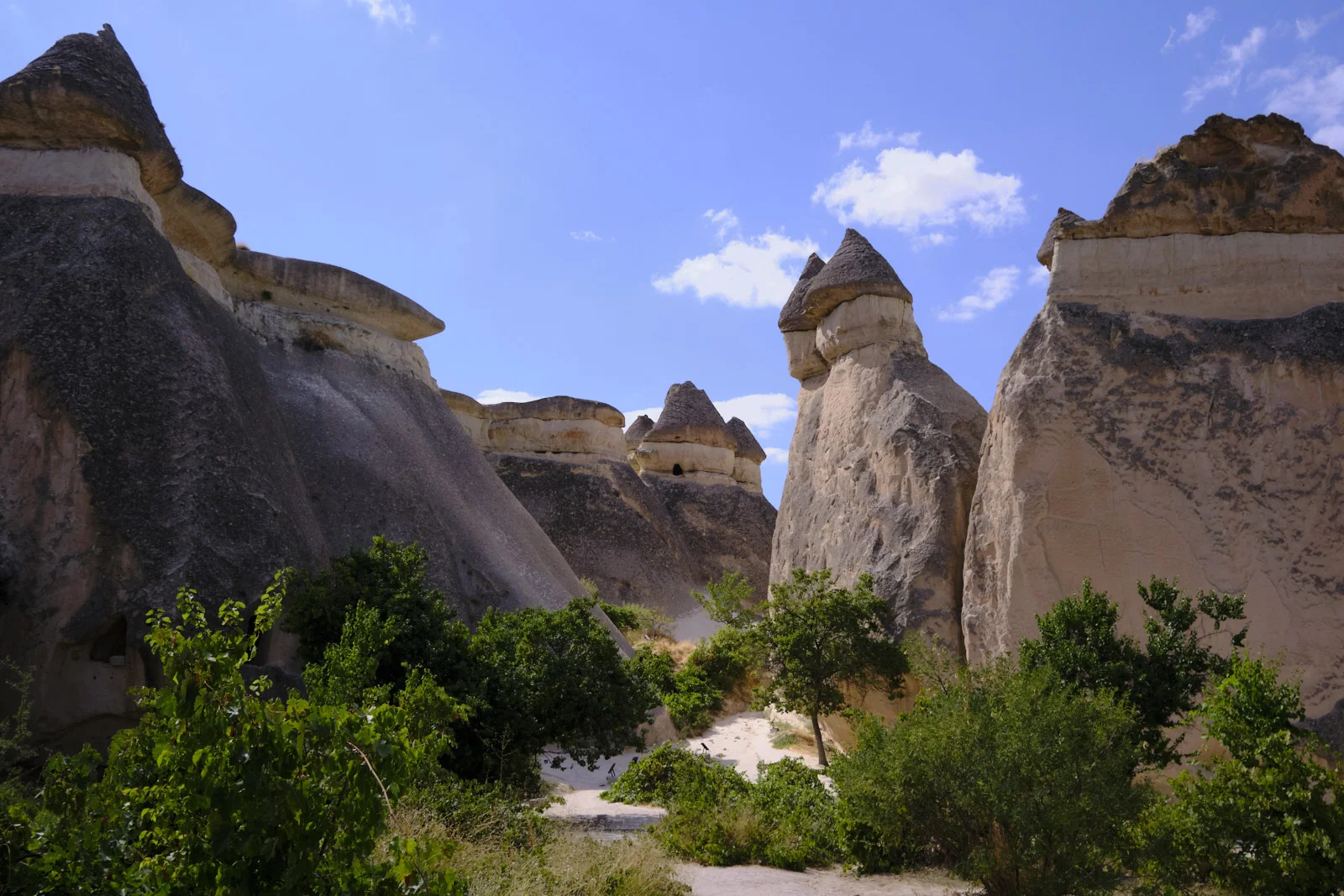 A sandy path passes between huge tuff cliffs toward a mushroom-capped fairy chimney under a blue sky with small clouds.