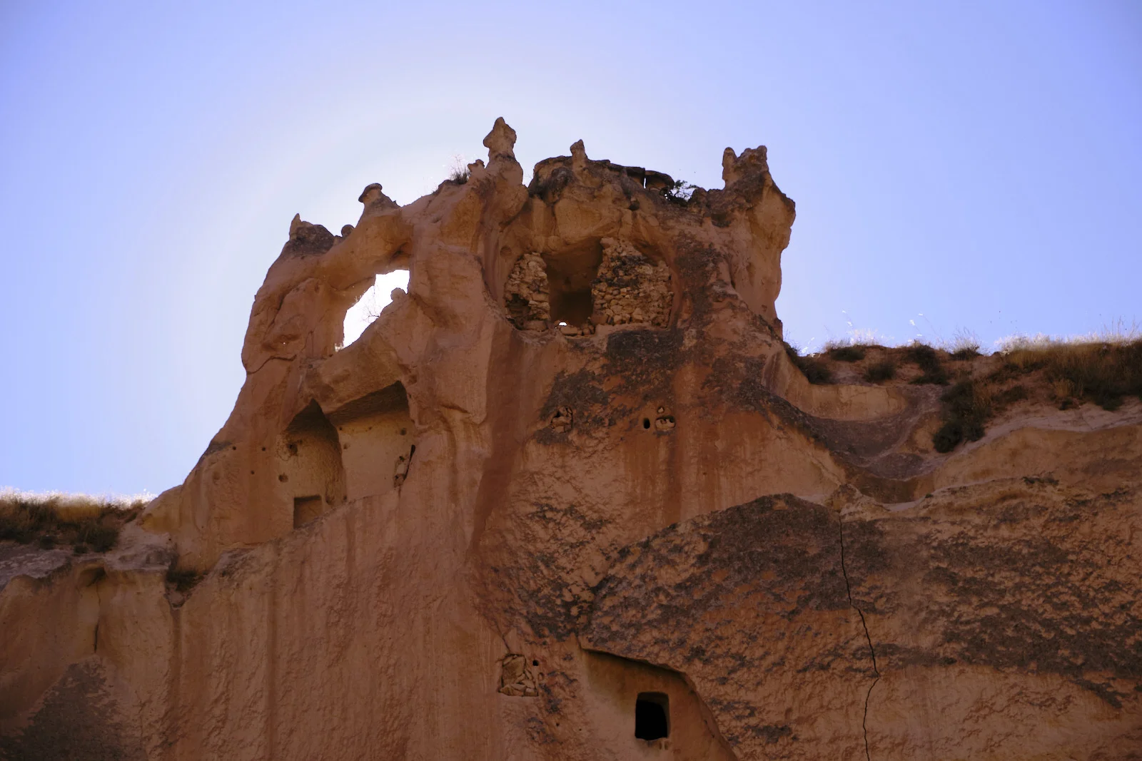 A sunlit tuff cliff pierced by dark cave openings, with stone-walled rooms near the top and a small doorway at the base.