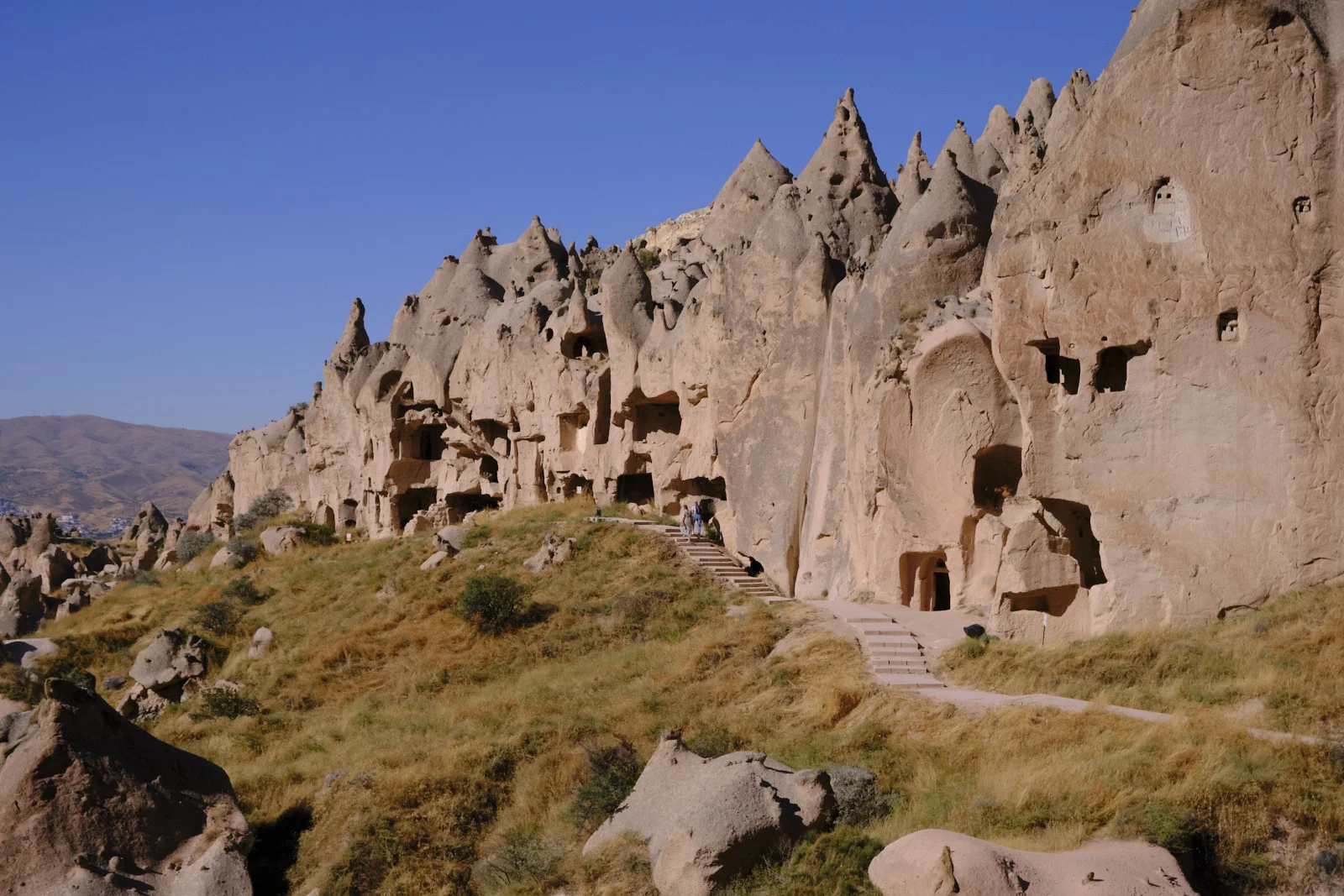 Visitors walk a path and stairs beneath a ridge of conical rocks honeycombed with dark cave openings.