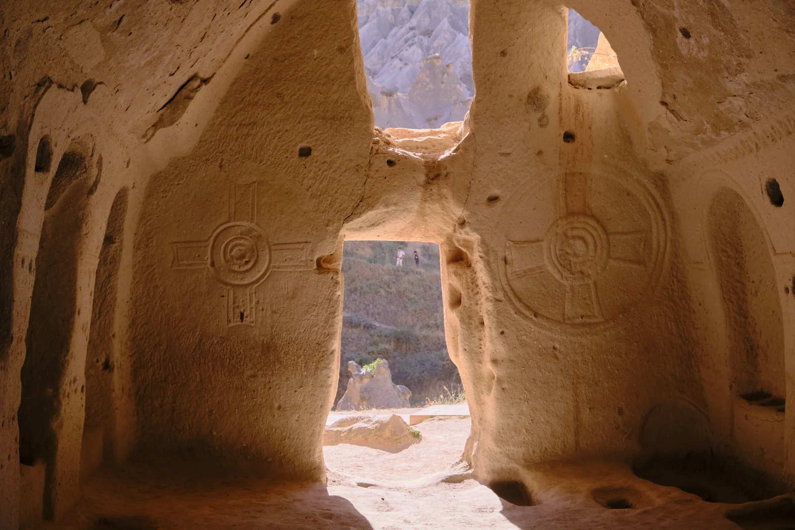 Carved stone crosses stand on either side of a rough doorway that opens onto a sunlit hillside and distant rock cones.