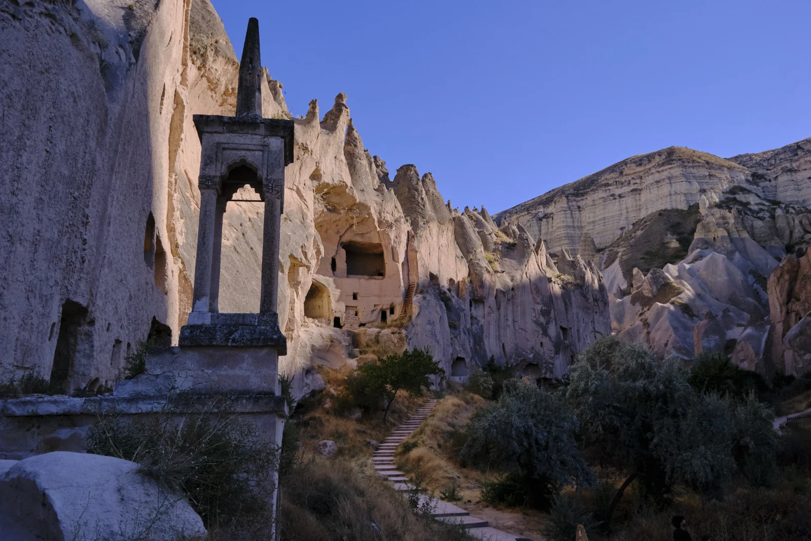A tall carved stone structure with an arched opening stands before a pale cliff of cave rooms, with steps winding through dry brush.