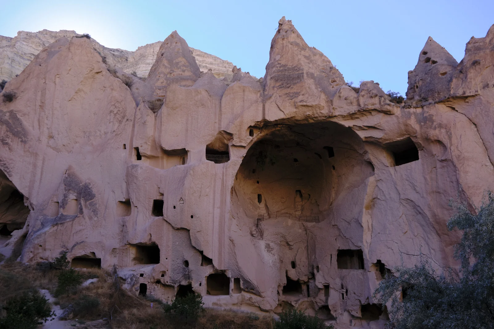 A massive pink-tan cliff is riddled with cave openings around one enormous central cavern, with walkways and dry brush at its base.