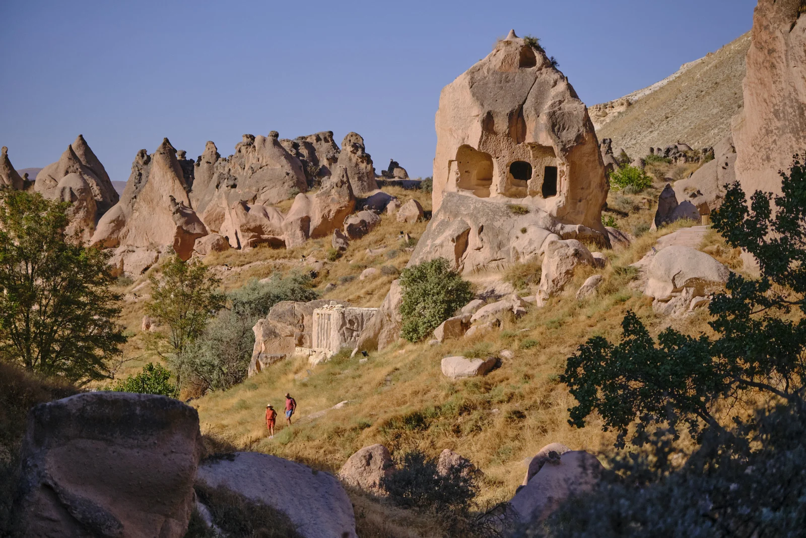 Two walkers pass below a large conical rock pierced by cave windows, with pointed formations and trees along the hillside.