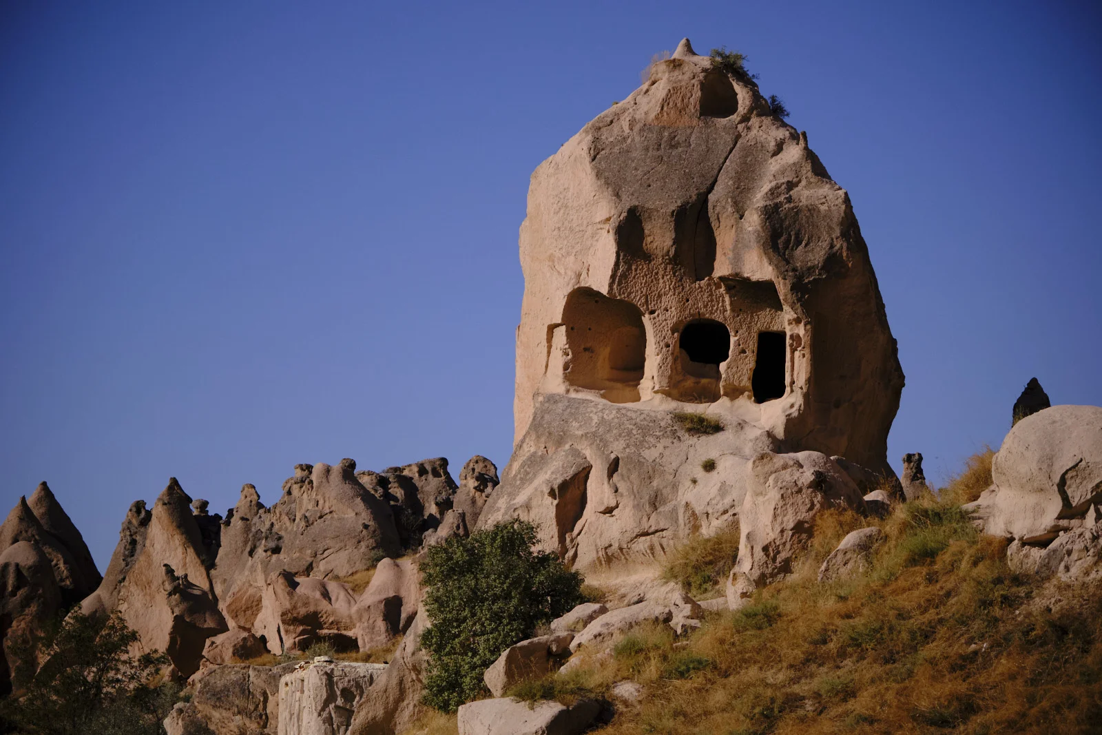 A tall conical rock with several dark window openings rises from a rocky, grass-covered slope against a clear blue sky.