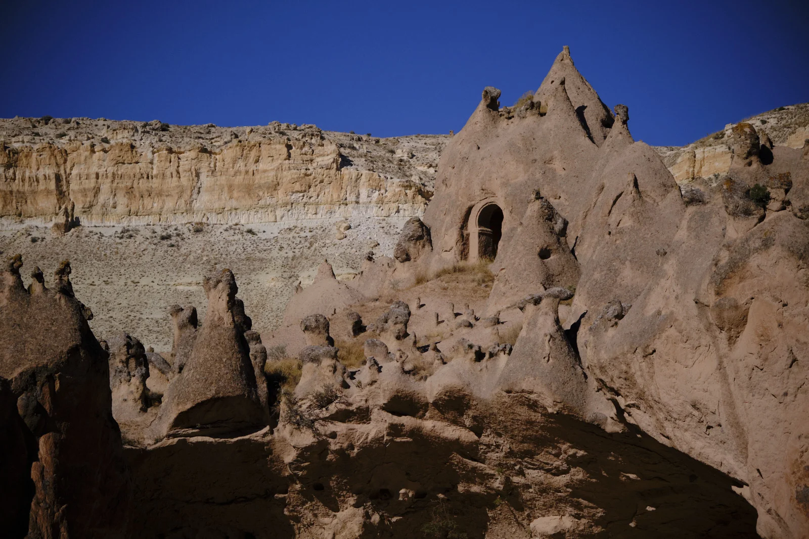 A wide view of eroded cones and a large rock with an arched opening, set against a striped cliff and deep blue sky.