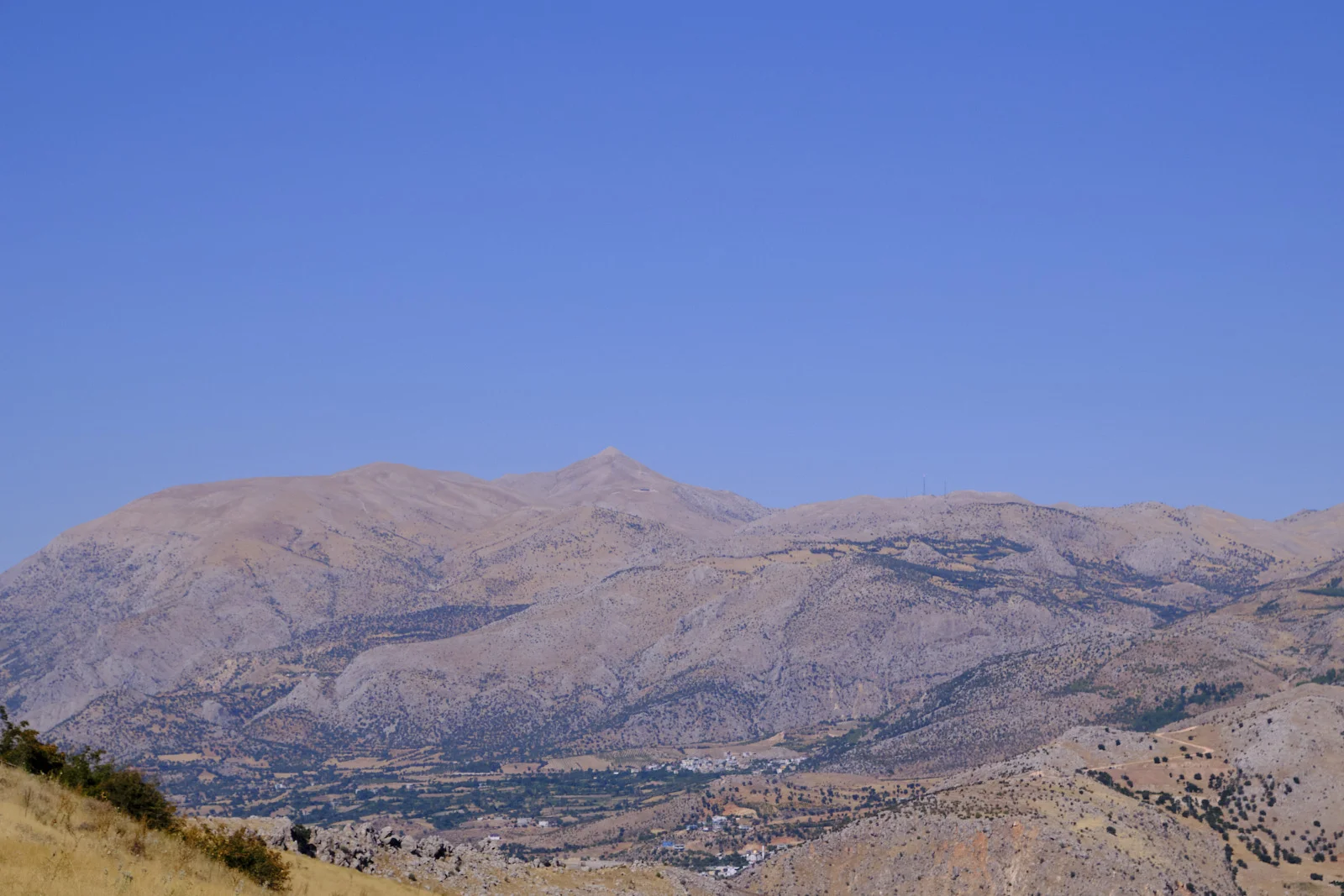A distant conical mountain peak rises above dry ridges and a valley settlement under a clear blue sky.