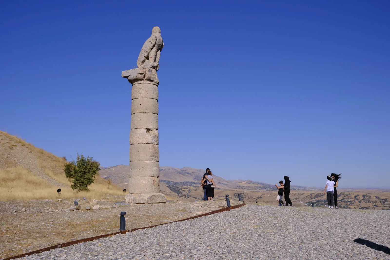 A stone eagle crowns a tall column on a gravel overlook, with visitors and distant mountains under a deep blue sky.