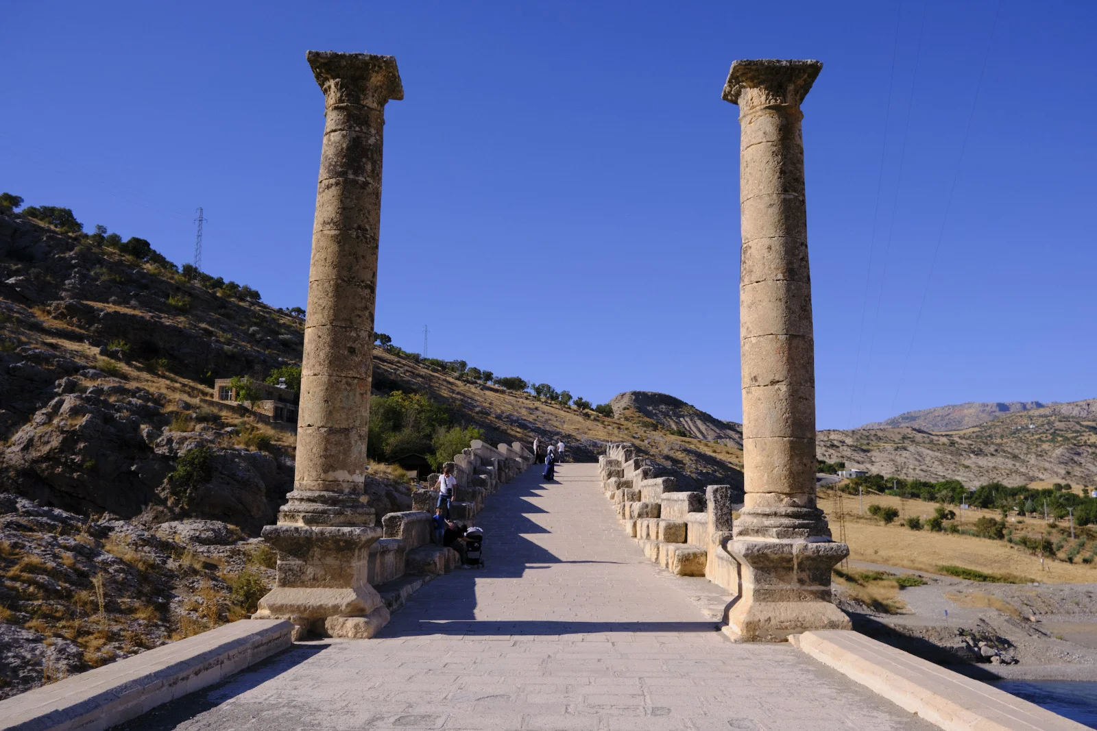 Two tall stone columns stand at the entrance of a paved Roman bridge, with visitors walking between the parapets.