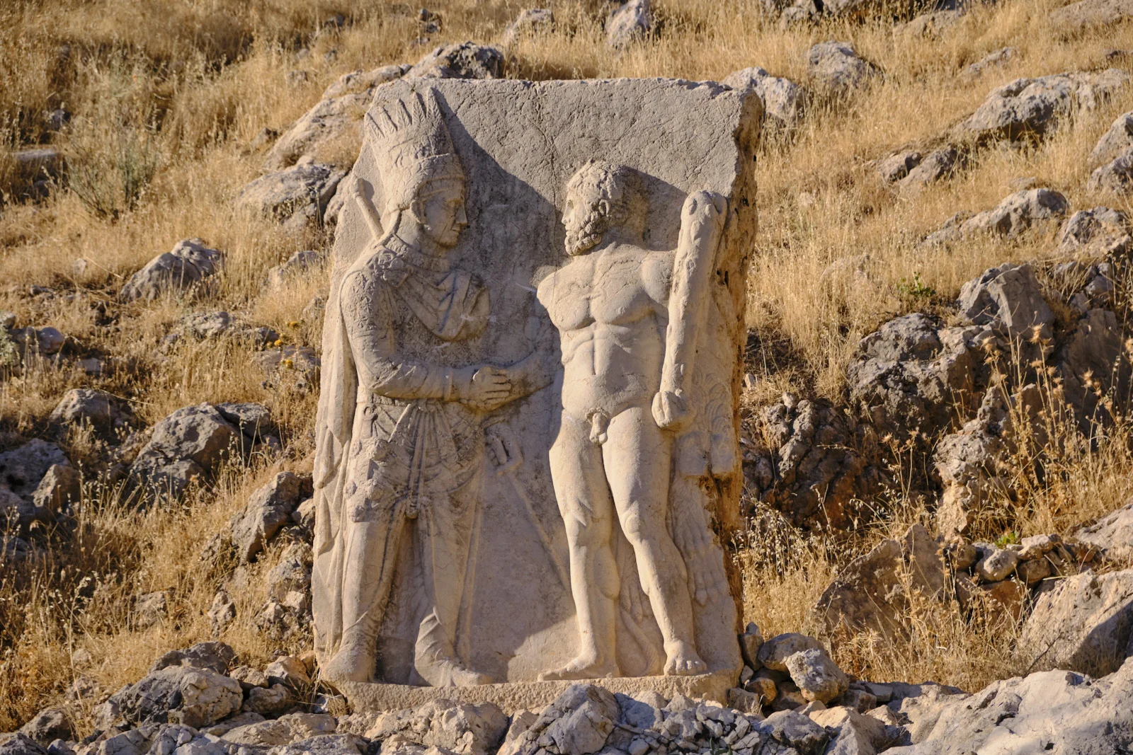 A stone relief shows a crowned king shaking hands with a nude, bearded Heracles holding a club, set among rocks and dry grass.