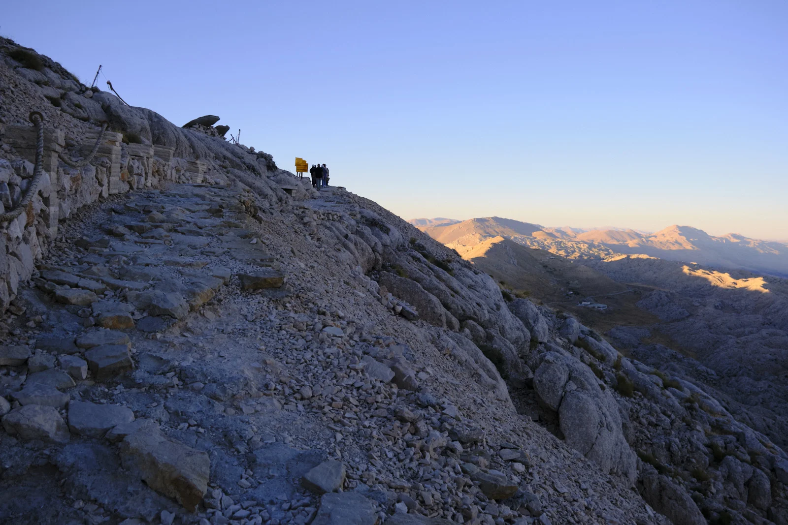 A rocky path and rope railing climb a mountain ridge where three people stand near yellow signs at dusk.