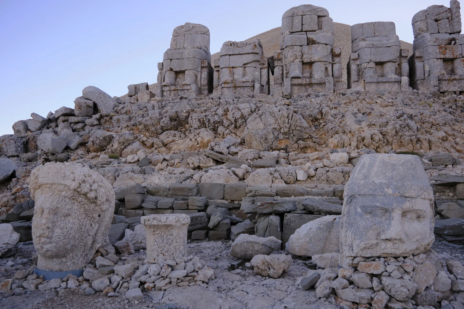 Two large stone heads lie among rubble beneath a row of headless seated statues on a rocky slope.