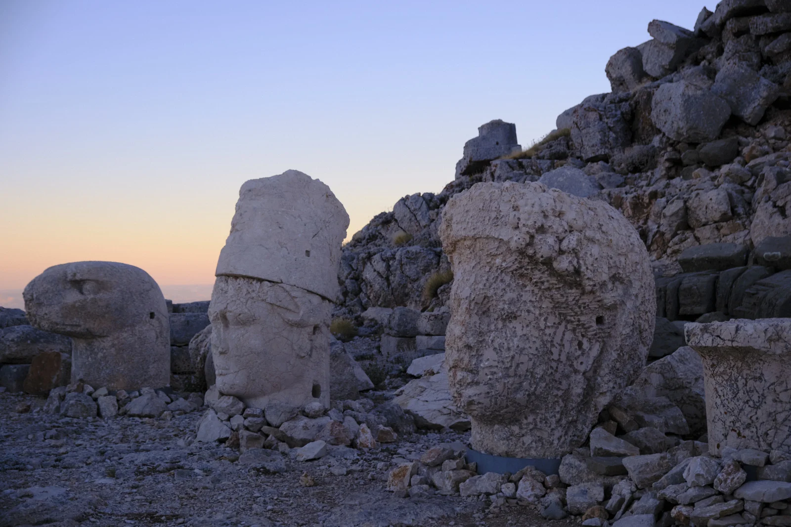 Three colossal stone heads, including an animal head and two human faces, stand among rocks against a fading sky.