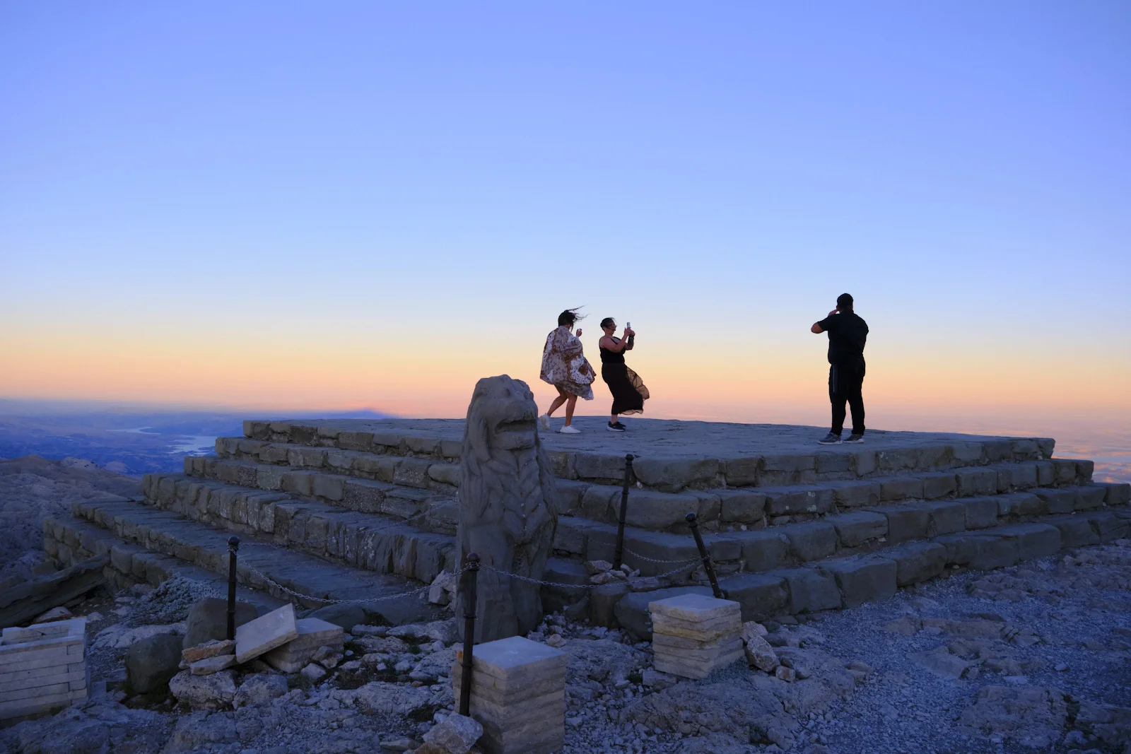 A carved stone lion stands at the corner of a summit platform where three people photograph the sunset.
