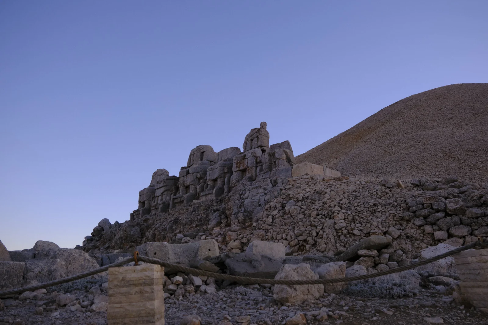 Headless block-built statues sit below a gravel burial mound, with a rope barrier in the foreground.