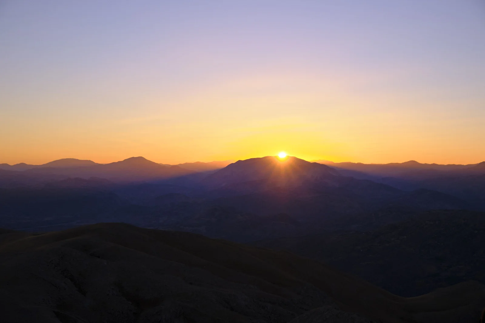 The sun sets behind a mountain ridge, lighting layered ranges in orange, gold, and deep blue.
